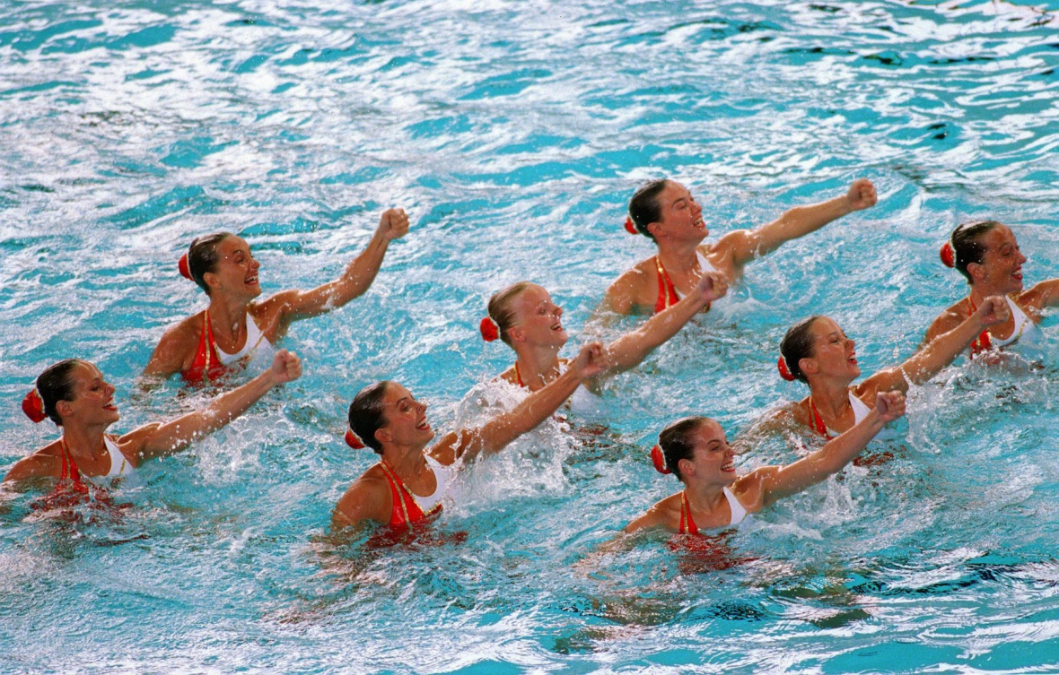 Huit nageuses pendant un programme en équipe sortent les épaules de l'eau et lèvent un bras.