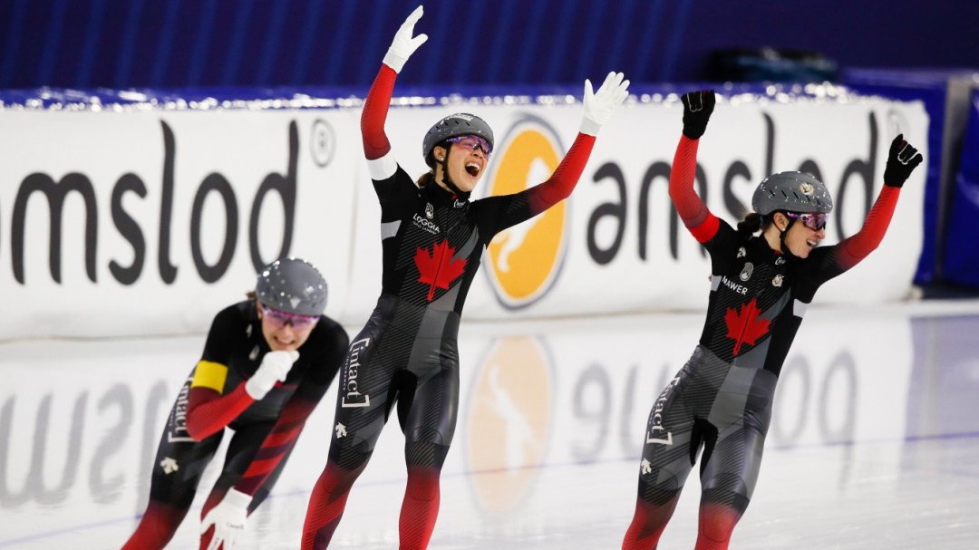 Trois patineuses de la poursuite par équipe d'Équipe Canada les bras levés vers le ciel célébrant leur victoire à la ligne d'arrivée.