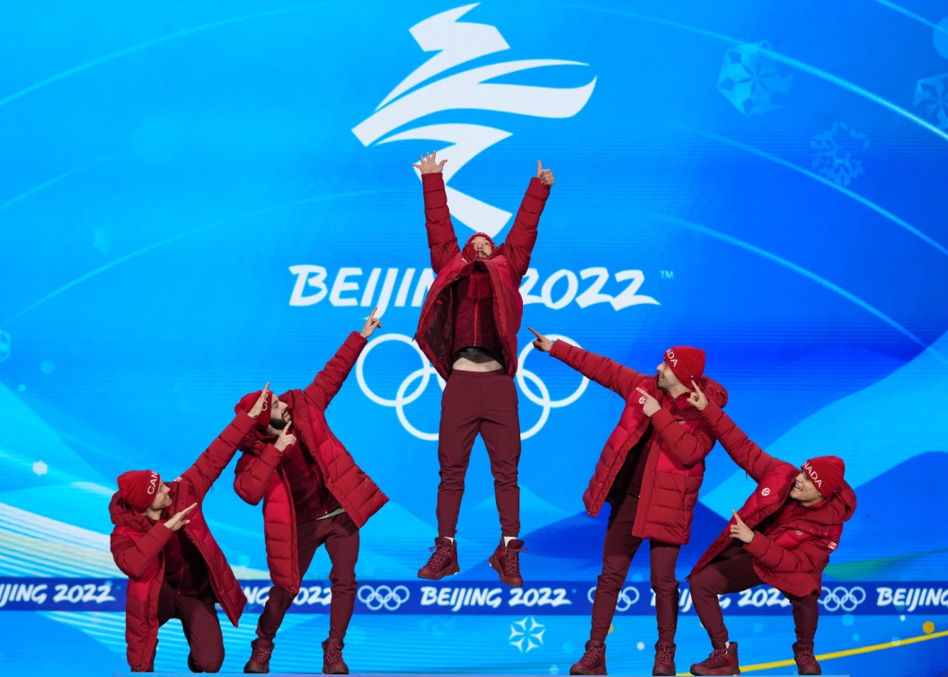 Charles Hamelin saute sur le podium alors que ses coéquipiers le pointent du doigt. Charles Hamelin est au centre tandis que deux coéquipiers sont de chaque côté, formant un triangle. Le logo des Jeux en blanc est sur un arrière-plan bleu.