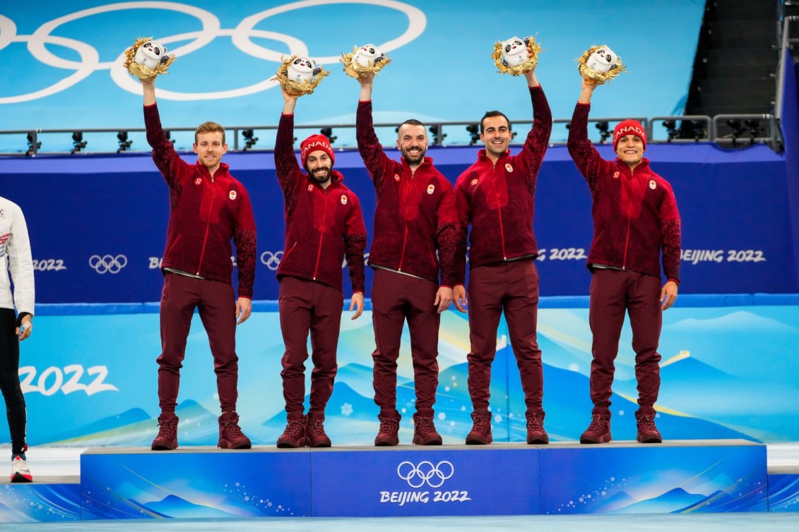 Pascal Dion, Steven Dubois,  Charles Hamelin, Maxime Laoun et Jordan Pierre-Gilles se tiennent debout, côte à côte sur le podium, et lèvent chacun une main vers le ciel avec la mascotte de Beijing 2022 pour célébrer leur médaille d'or gagnée au relais de 5000 mètres. 