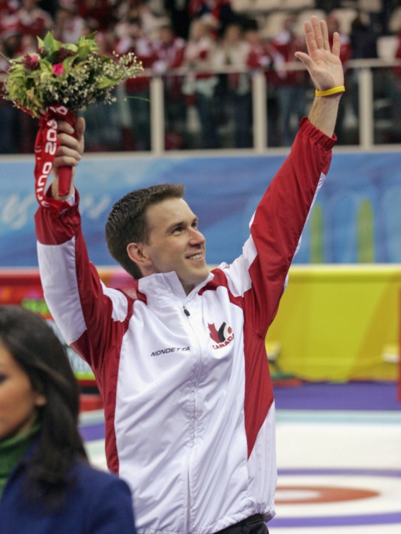 L'athlète canadien de curling Brad Gushue lève les bras pour saluer la foule en tenant un bouquet de fleurs dans sa main droite. 