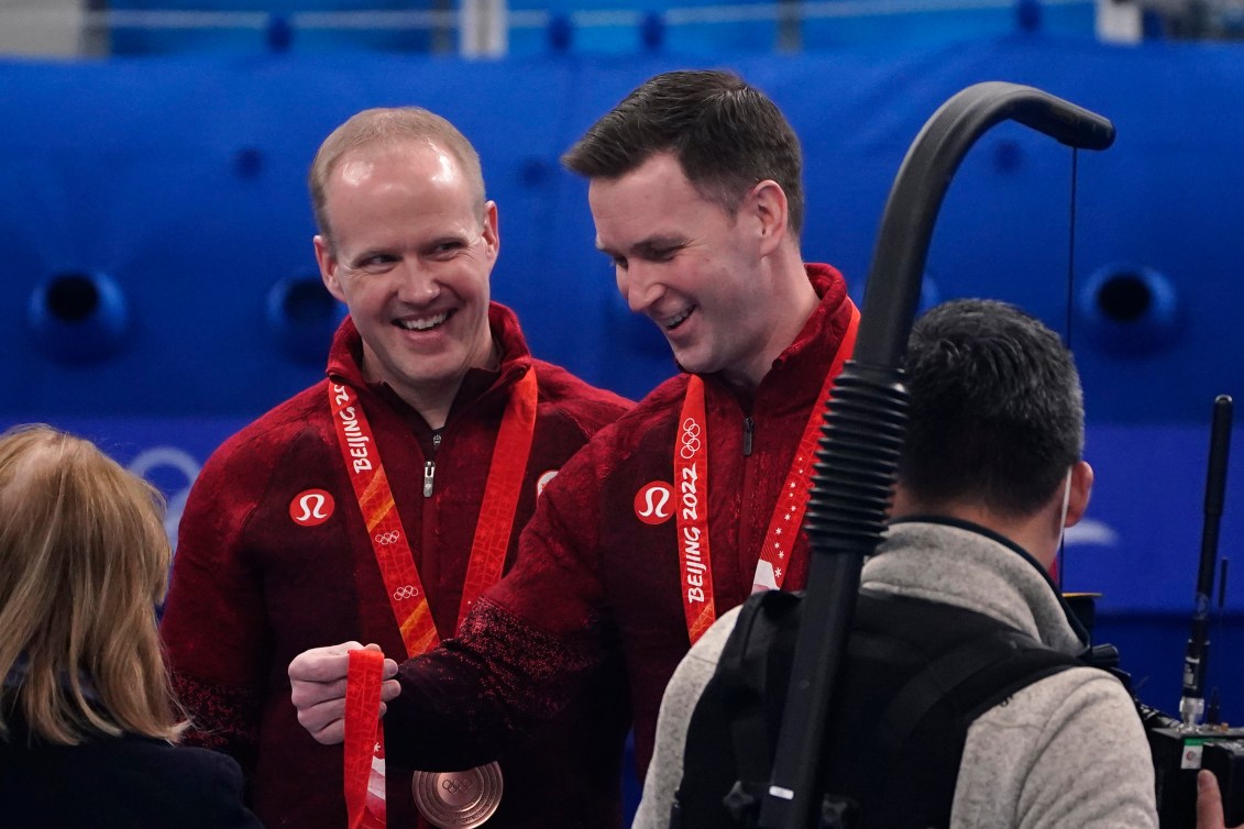 Le Canadien Brad Gushue, à droite, regarde une médaille de bronze avec son coéquipier Mark Nichols à ses côtés. Ils portent tous deux leur médaille de bronze au ruban rouge au cou.