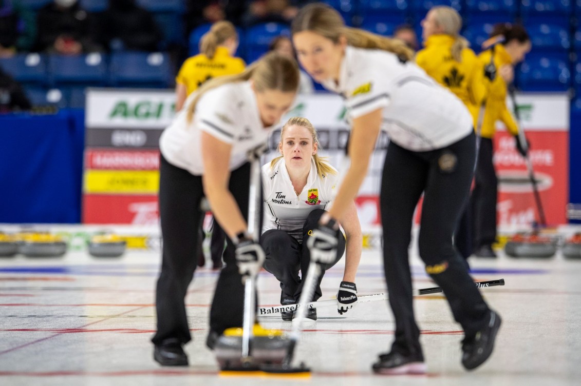 Trois joueuses de curling sur la surface. Une vient de lancer et deux balayent.