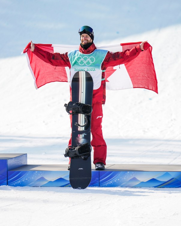 Le planchiste d'Équipe Canada Max Parrot fait voler le drapeau candien dans son dos alors qu'il est derrière et sa planche et sur le podium du big air en snowboard masculin à Beijing 2022.