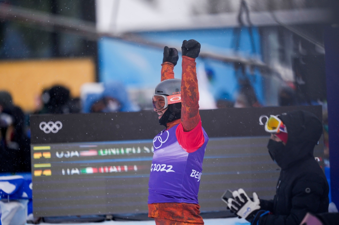 Eliot Grondin lève les bras dans les airs quand il aperçoit Meryeta O’Dine arriver à la ligne d'arrivée. Il porte un dossard mauve par-dessus ses vpetements de skis rouges. Il porte ses lunettes de ski.