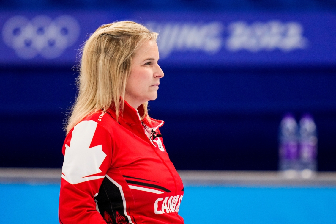 Jennifer Jones est en avant-plan debout de côté, regardant au loin. Elle a un regard neutre et porte sa veste rouge à l'effigie d'Équipe Canada.. On aperçoit en arrière-plan les anneaux olympiques et la signature Beijing 2022 embrouillé sur un mur bleu.