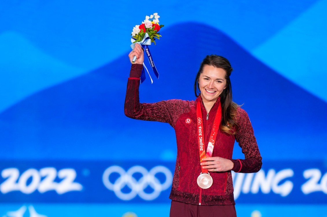 La patineuse canadienne longue piste Isabelle Weidemann sur le podium du 3000 m féminin à Beijing 2022. Elle porte sa médaille de bronze autour du cou et lève le bras droit avec le bouquet de fleurs. 