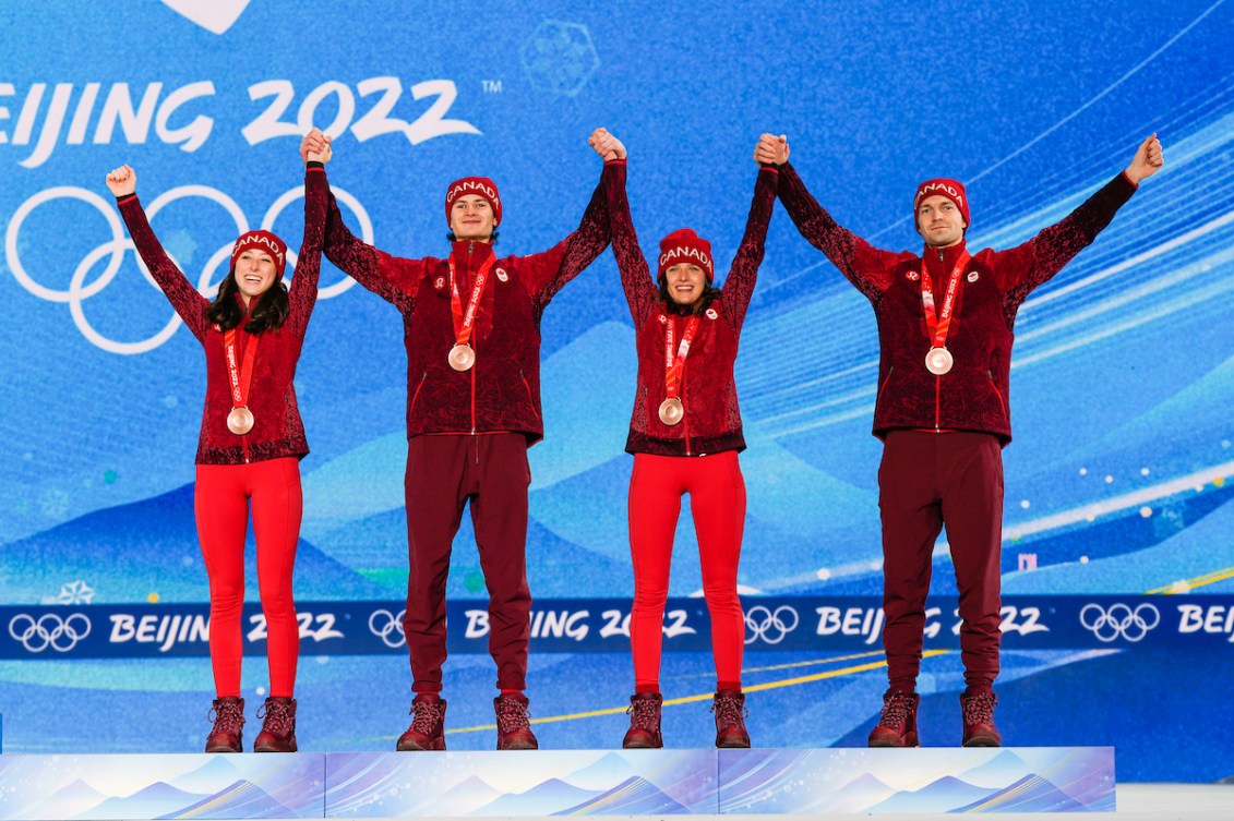 Les membres de l'équipe canadienne du saut à ski par équipes mixtes, Alexandria Loutitt, Matthew Soukup, Abigail Strate et Mackenzie Boyd-Clowes se tiennent les mains et lèvent celles-ci dans les airs en portant leur médaille de bronze autour du cou sur le podium de l'épreuve aux Jeux de Beijing 2022.
