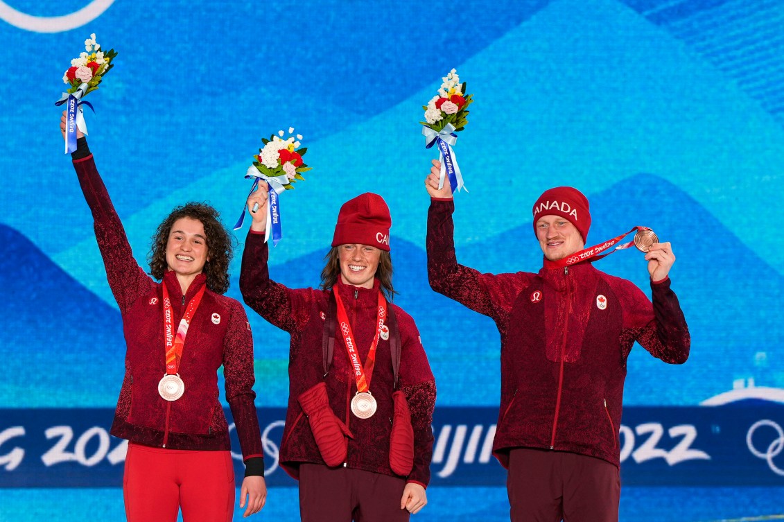 Les skieurs acrobatiques d'Équipe Canada Marion Thénault, Miha Fontaine et Lewis Irving soulèvent leur bouquet de fleurs et Lewis Irving soulève sa médaille de bronze sur le podium du saut acrobatique mixte par équipes à Beijing 2022.