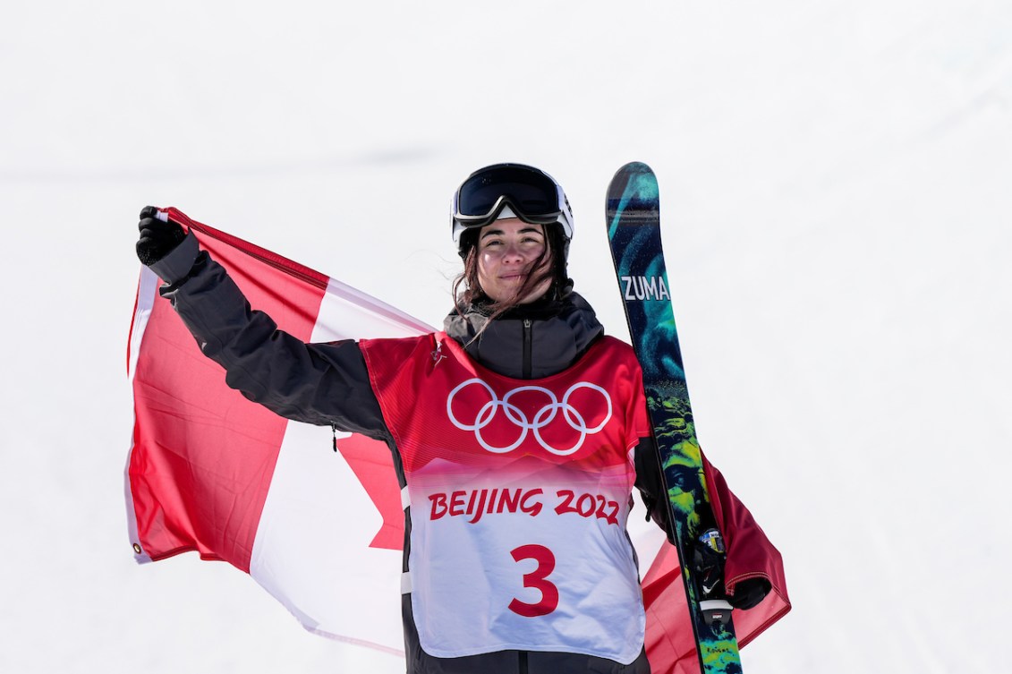 La skieuse acrobatique d'Équipe Canada Rachael Karker porte le drapeau canadien dans son dos et ses skis de la main gauche après sa médaille de bronze à la demi-lune en ski acrobatique à Beijing 2022.