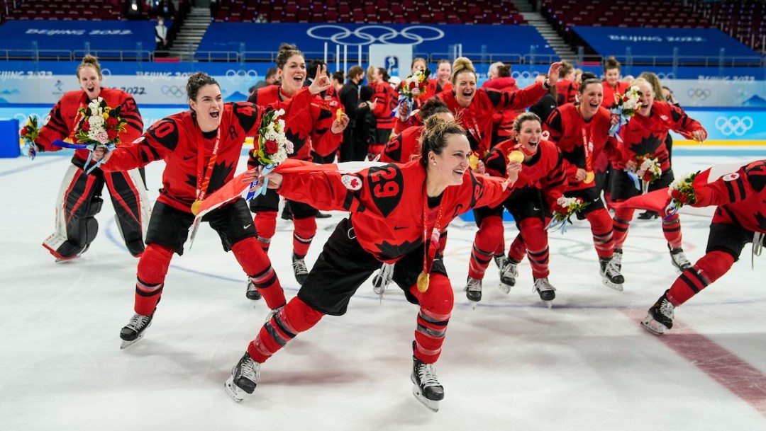 En avant-plan, Marie-Philip Poulin tient le drapeau canadien dans son dos comme une cape. En arrière-plan, le reste d'Équipe Canada célèbre la victoire en hockey féminin à Beijing 2022 avec sa médaille d'or autour du cou.