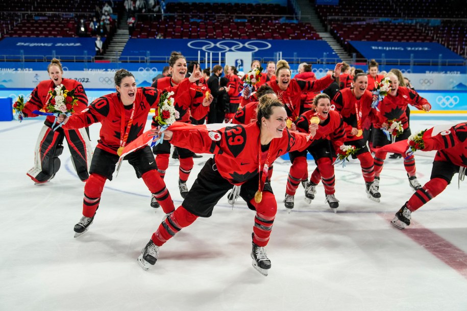 En avant-plan, Marie-Philip Poulin tient le drapeau canadien dans son dos comme une cape. En arrière-plan, le reste d'Équipe Canada célèbre la victoire en hockey féminin à Beijing 2022 avec sa médaille d'or autour du cou.