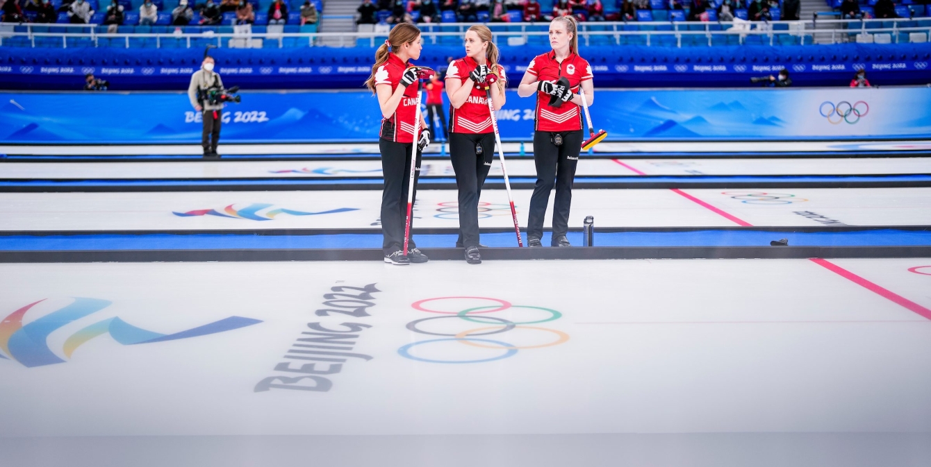 Trois joueuses de curling d'Équipe Canada se tiennent debout sur la glace. En avant-plan, le logo des Jeux olympiques d'hiver de Beijing est imprégné dans la glace.