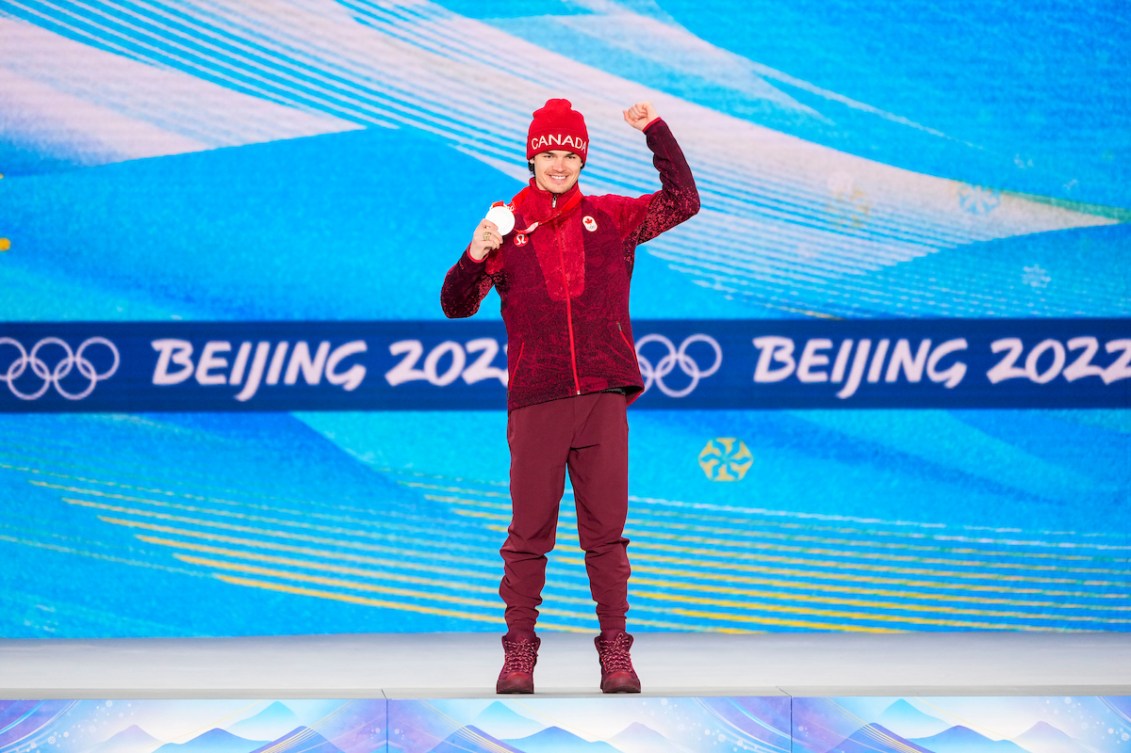 Mikaël Kingsbury d'Équipe Canada tient sa médaille d'argent de l'épreuve des bosses en ski acrobatique avec sa main droite et soulève le bras droit sur le podium aux Jeux olympiques d'hiver de Beijing 2022.