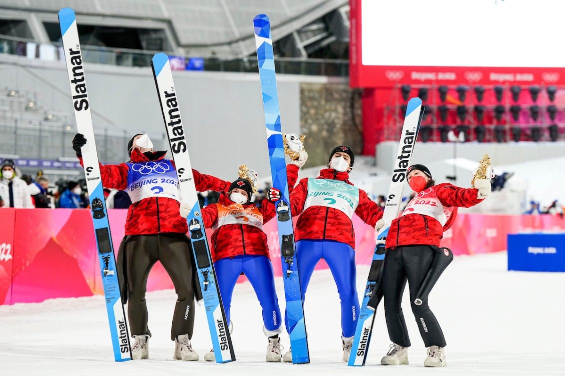 Les sauteurs à ski d'Équipe Canada Abigail Strate, Matthew Soukup, Alexandria Loutitt et Mackenzie Boyd-Clowes tiennent leurs skis de la main droite, la mascotte des Jeux de la main gauche et ils se penchent vers l'arrière pour célébrer leur médaille de bronze à l'épreuve de saut à ski mixte par équipes à Beijing 2022.