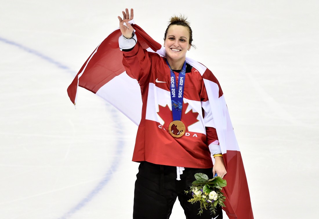 Marie-Philip Poulin sur la glace avec la médaille d'or de Sotchi autour du cou, un bouquet de fleurs dans la main gauche, et le drapeau canadien sur les épaules, salue de la main droite.