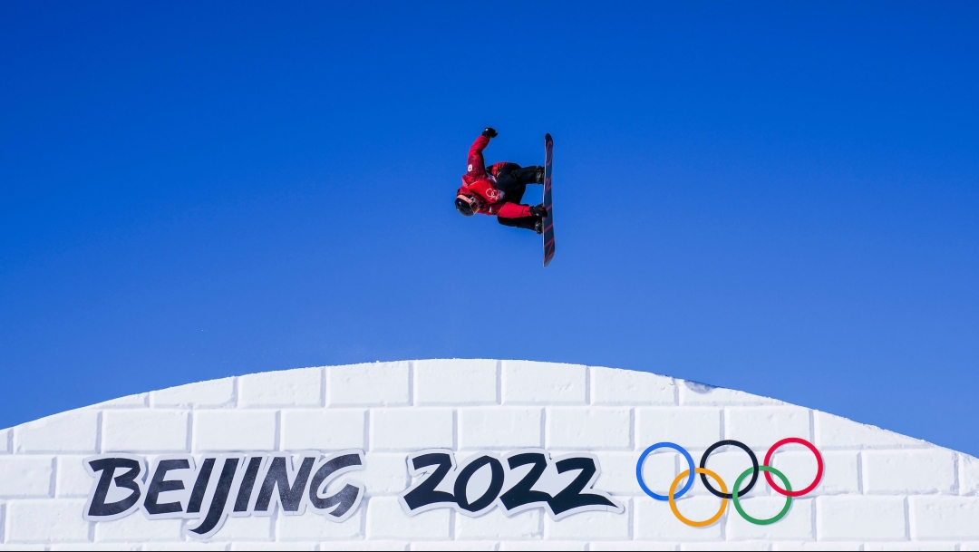 Le planchiste Mark McMorris en plein saut, sa planche perpenticulaire au sol sur un fond de ciel complètement bleu. Il a un manteau rouge et des pantalons noir. Il est au-dessus d'une pente blanche de neige sur laquelle la signature visuelle Beijing 2022 est inscrite en noir avec les anneaux olympiques de couleur à droite.