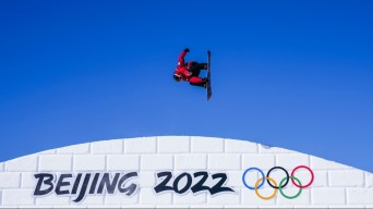 Le planchiste Mark McMorris en plein saut, sa planche perpenticulaire au sol sur un fond de ciel complètement bleu. Il a un manteau rouge et des pantalons noir. Il est au-dessus d'une pente blanche de neige sur laquelle la signature visuelle Beijing 2022 est inscrite en noir avec les anneaux olympiques de couleur à droite.