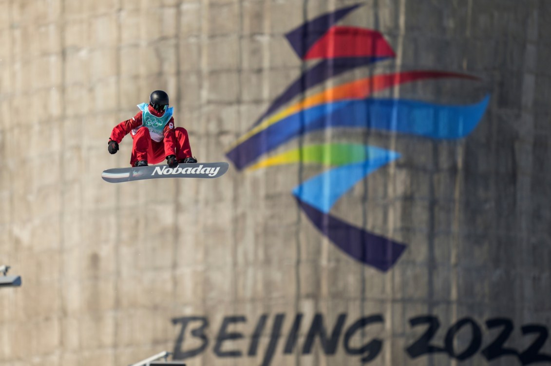 Le planchiste d'Équipe Canada Max Parrot prend sa planche avec sa main gauche en faisant une figure dans les airs au concours de big air de Beijing 2022.