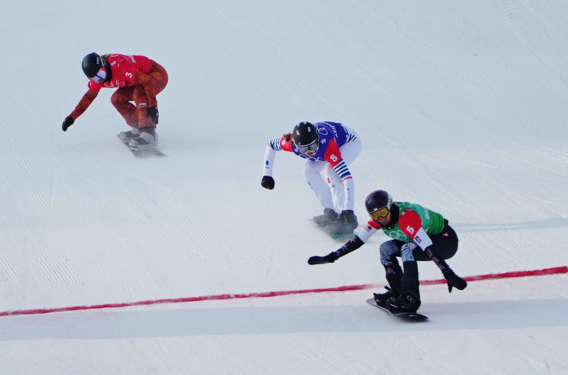 Trois planchistes pendant une course de snowboard cross. Le dossard vers est devant, suivi de près par le dossard bleu et le dossard rouge quelques mètres derrière. 