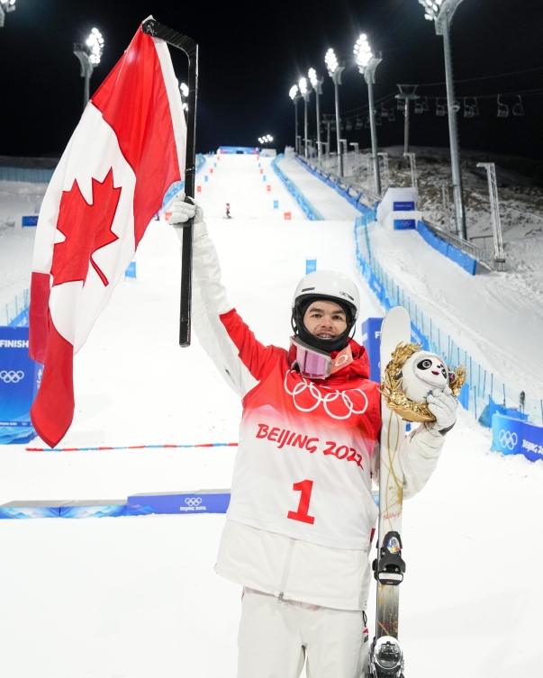 Mikaël Kingsbury sourit avec le drapeau canadien dans la main droite et une peluche de la mascotte de Beijing 2022 dans la main gauche.