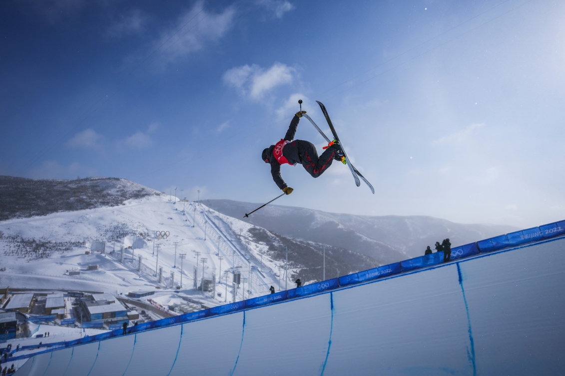 Noah Bowman d'Équipe Canada exécute un tour de ski dans l'épreuve de demi-lune. Il est dans les airs, au-dessus de la rampe avec ses akis croisés. Au loin, on voit les montagnes enneigées et la piste de ski alpin.