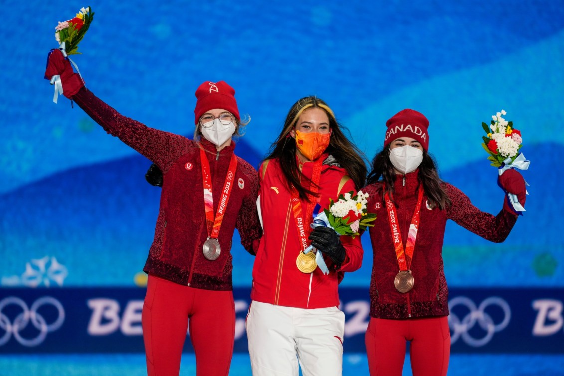 Les skieuses acrobatiques d'Équipe Canada Cassie Sharpe, à gauche, et Rachael Karker, à droite entourent la championne olympique Eileen Gu sur le podium de demi-lune en ski acrobatique.