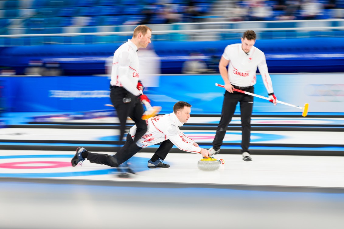 Le capitaine d'Équipe Canada, Brad Gushue, lance la pierre contre l'équipe de Norvège lors du tournoi à la ronde lors des Jeux olympiques d'hiver de Beijing 2022. Pendant qu'il est au sol, deux coéquipiers sont debout à côté de lui avec balais en main.