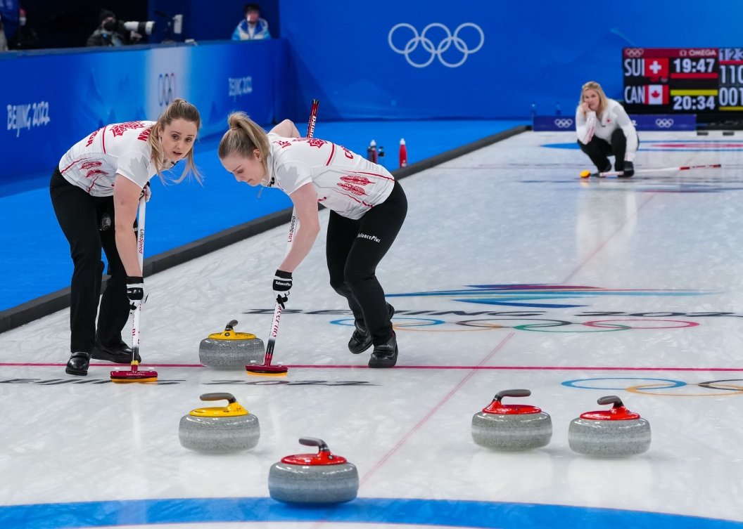 Jennifer Jones regarde son tir de la pierre au curling être balayé par Jocelyn Peterman et Dawn McEwen. Jennifer Jones est au loin sur la glace accroupie alors que ses coéquipières sont en avant-plan. Elles balyent la pierre en mouvement jaune. On aperçoit trois pierres rouges et une jaune en avant-plan.