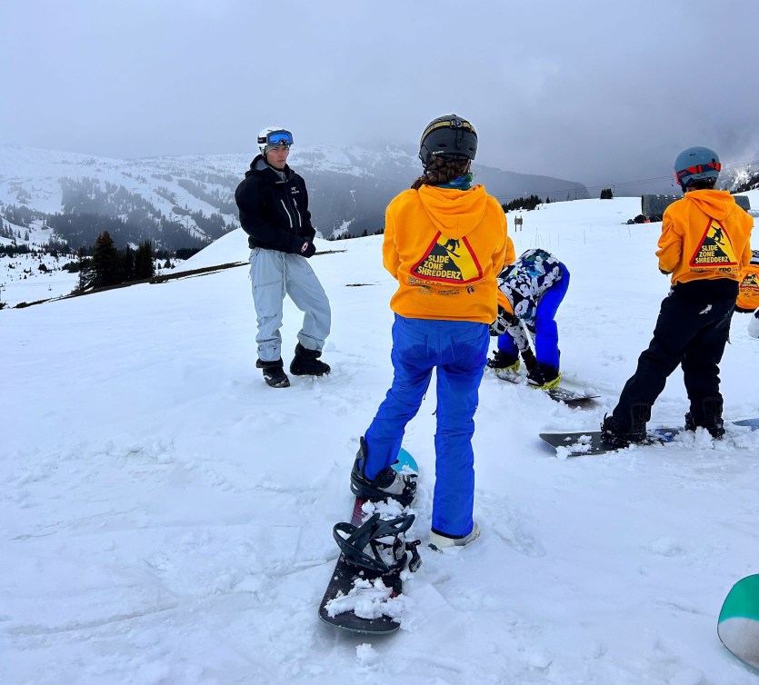 Liam Gill en train de donner des instructions à de jeunes planchistes autochotones, à l'extérieur, sur une montagne enneigée.