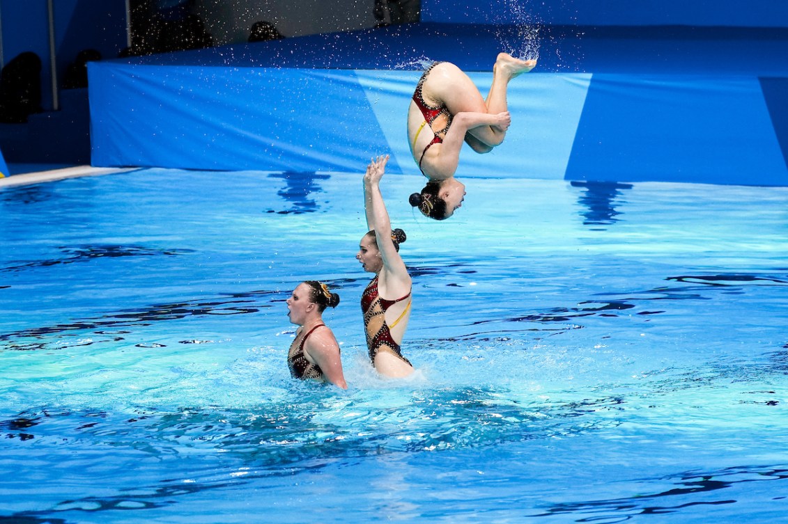 Trois nageuses canadiennes, dont celle du milieu qui en propusle une autre derrière elle.  Cette dernière fait une pirouette dans les airs.