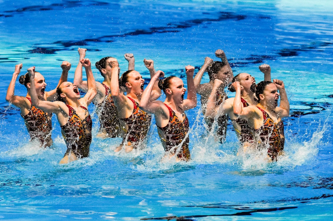 L'ensemble de l'équipe canadienne de natation artistique en train de performer dans la pisicine.  Elles ont toutes les bras relevés, pliés vers la tête.