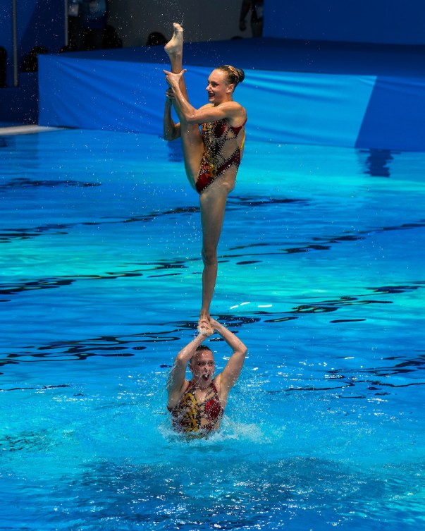 Une nageuse artistique exécute un équilibre sur une jambe soutenue par une coéquipière. 