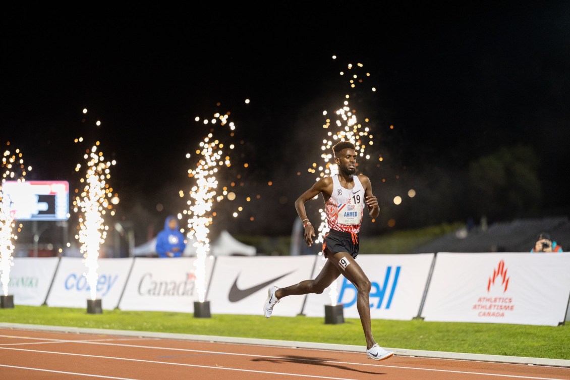 Mo Ahmed, entrain de courir sur la piste d'athlétisme, avec des petits feux d'artifices derrière lui.