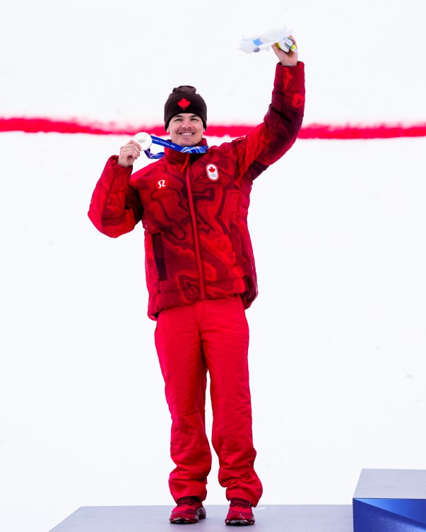 Mikaël Kingsbury célèbre sur le podium avec sa médaille dans une main et une peluche de la mascotte dans l'autre. 